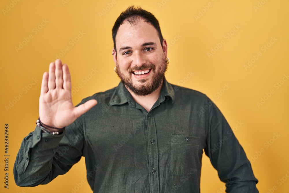 Plus size hispanic man with beard standing over yellow background waiving saying hello happy and smiling, friendly welcome gesture Plus size hispanic man with beard standing over yellow background waiving saying hello happy and smiling, friendly welcome gesture
