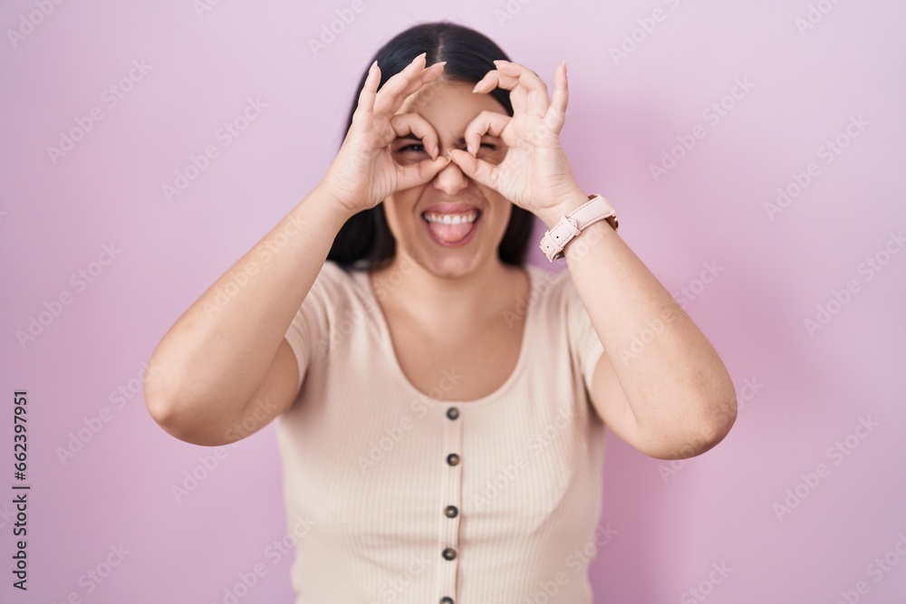 Young hispanic woman standing over pink background doing ok gesture like binoculars sticking tongue out, eyes looking through fingers. crazy expression. Young hispanic woman standing over pink background doing ok gesture like binoculars sticking tongue out, eyes looking through fingers. crazy expression.