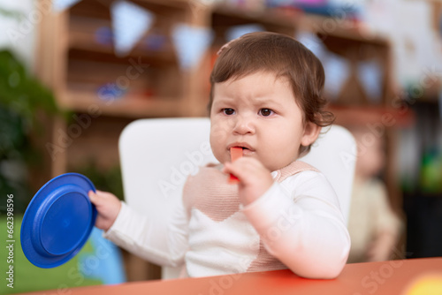 Adorable toddler sitting on table sucking plastic spoon holding dish at kindergarten