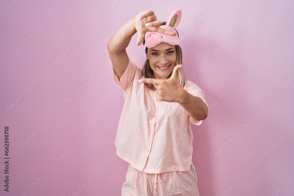 Blonde caucasian woman wearing sleep mask and pajama smiling making frame with hands and fingers with happy face. creativity and photography concept.
