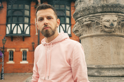 Welcome concept. Young bearded romantic man wearing pink trendy hoodie posing over brick house with flowers on backgound. Old classical fontain. Close up. Outdoor shot photo