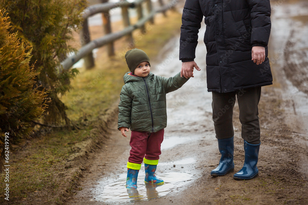 Father and son walking in the fresh air in rubber boots on the puddles and mud after the rain. Little child holding hand of a man Father and son walking in the fresh air in rubber boots on the puddles and mud after the rain. Little child holding hand of a man