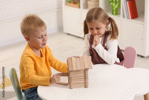 Little boy and girl playing with wooden house at white table indoors. Children's toys