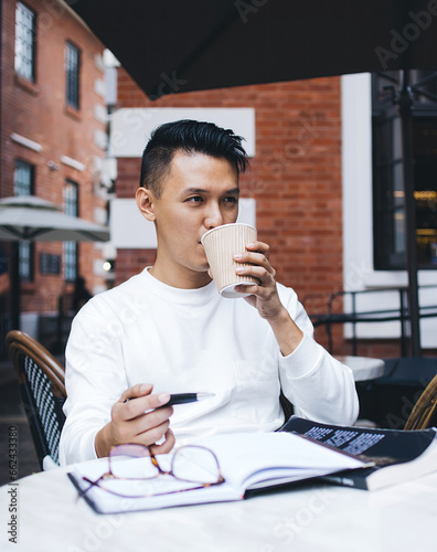 Adult ethnic man having hot drink and contemplating while sitting at table on cafe terrace