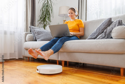 Robotic vacuum cleaner cleaning the living room while woman sitting on sofa