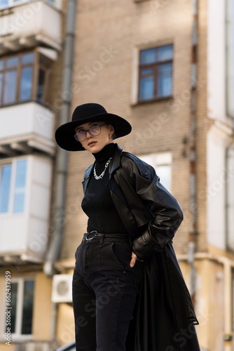 Portrait of an attractive and young blonde woman in a black hat and a leather trench coat while walking around the city. The concept of style and fashion