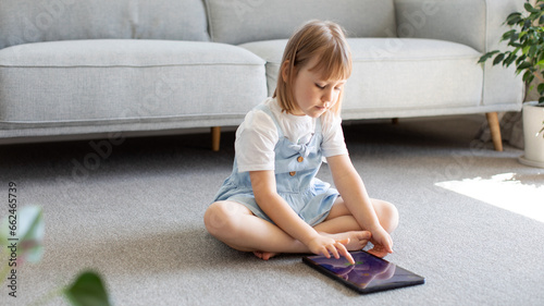 A cute girl looks at the tablet screen while sitting on the floor in the room. Children's educational apps for tablet. 