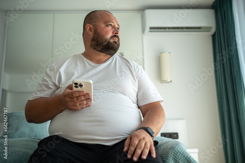 Obese unshaved man with phone and fitness tracker looking at window. Overweight middle-aged man thinks on weight reduction, diet and calories calculation. Weight control and healthy food concept photo