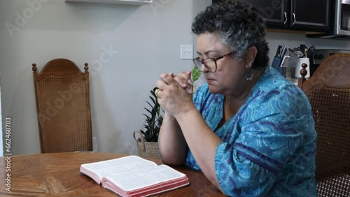 Senior woman with Gray curly hair is praying at kitchen table while having Bible open for reading