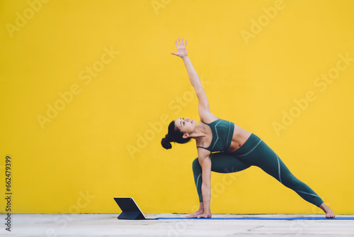 Young sporty fit woman exercising yoga in revolved side angle pose using tablet in yard