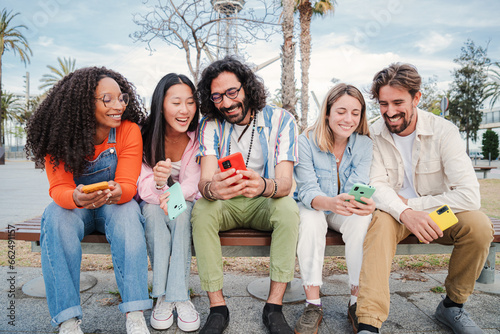 Group of multiracial young adult friends using their cellphones, browsing on internet enjoying free time sitting on bench outside. Encounter of happy people having fun together with their smart phones