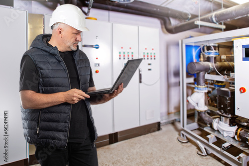 Man is industrialist. Guy in production room. Engineer holding laptop. Man works in industrial enterprise. Engineer controls equipment via computer. Factory technologist in white hardhat