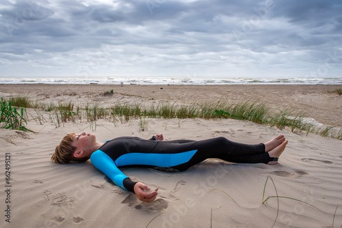 Young boy in a wet suit lying down resting on a sand dune at the beach