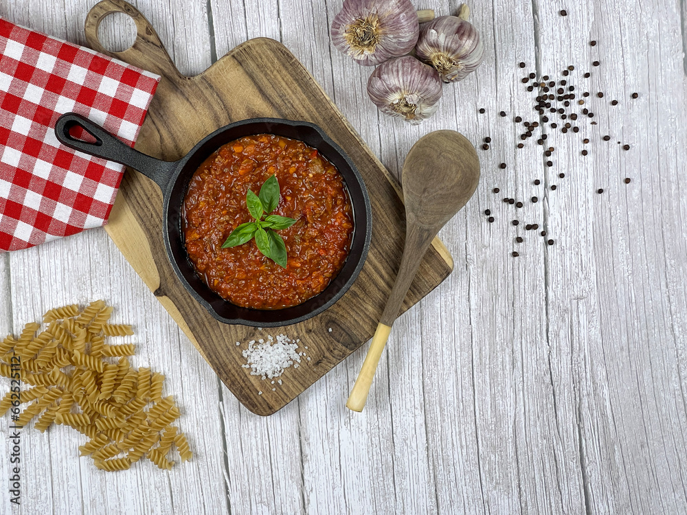 Spaghetti sauce in a cast iron skillet, pastas, garlic, and peppercorns, red and white checked fabric napkin on a wooden background, top view, copy space