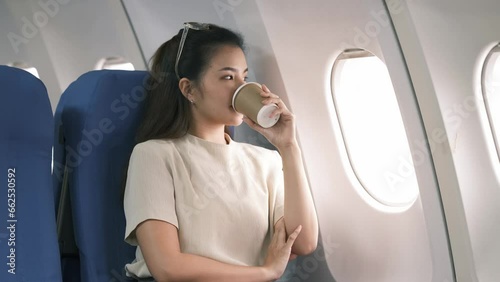 Asian woman airplane passenger with coffee cup looking at plane window, woman traveler in casual wear sitting in a plane.