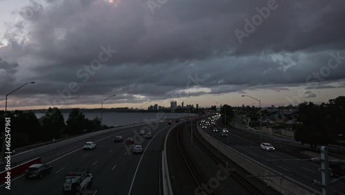 Very wide angle view of freeway traffic at dusk. Train line between lanes. Perth City, Swan River in background. Grey clouds with purple tinges fly overhead in timelapse. Bright car lights flash past