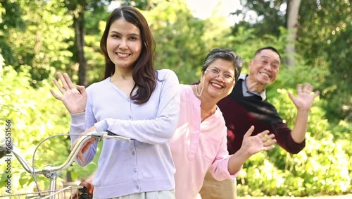 Asian family parents and their grown daughter stood with their bicycle handlebars in line greeting each other by waving and hiding behind them gazing into the camera smiling happily in the garden. photo
