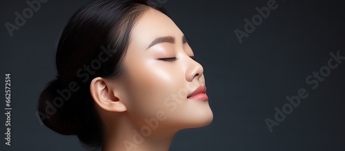 Asian woman with brown hair in ponytail touching her shoulder eyes closed focused on skincare With copyspace for text