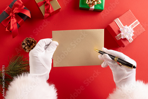 Santa claus holding envelope and pencil with copy space and christmas presents on red background