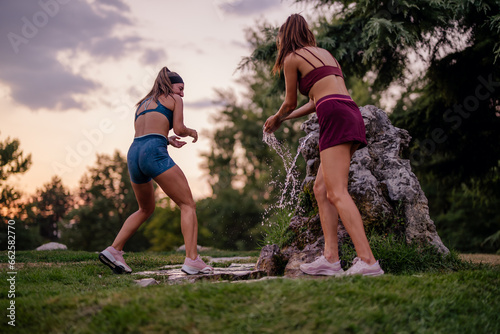 Fit, muscular women having fun, splashing water on each other after training in a green park. Their athletic bodies and happy smiles showcase an active and healthy lifestyle.