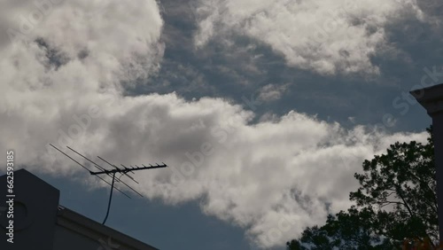 Strange wind patterns stir clouds to move in unusual opposite directions as they sweep above large tree and roofline of suburban houses. Timelapse
