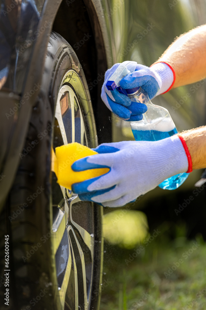 Man in protective gloves holding sponge and spray bottle while cleaning car rims outdoor