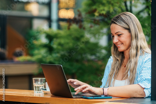Seated in the café's outdoor area, a woman in her thirties, a freelancer, diligently works on her laptop, surrounded by the garden's tranquility.