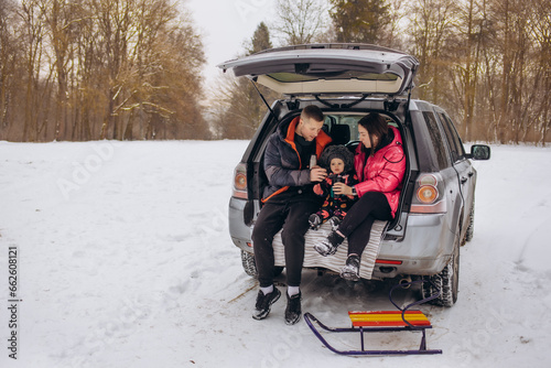 Happy family with dog on vacation during the winter holidays near road. Dressed in warm clothes sitting on the trunk of a car and drinking tea from a thermos. space for text. Winter vacation