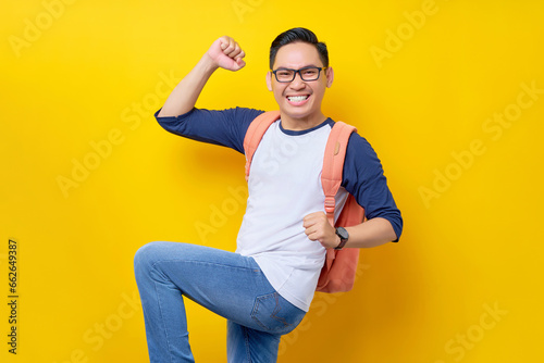 Excited young Asian student man in casual clothes and glasses backpack standing confident while celebrating success isolated on yellow background. Education in High School University College concept