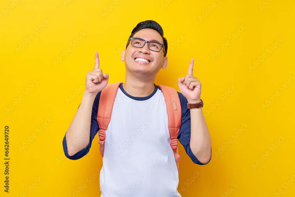 Smiling young Asian student man in casual clothes and glasses backpack pointing finger up at empty space isolated on yellow background. Education in High School University College concept Smiling young Asian student man in casual clothes and glasses backpack pointing finger up at empty space isolated on yellow background. Education in High School University College concept