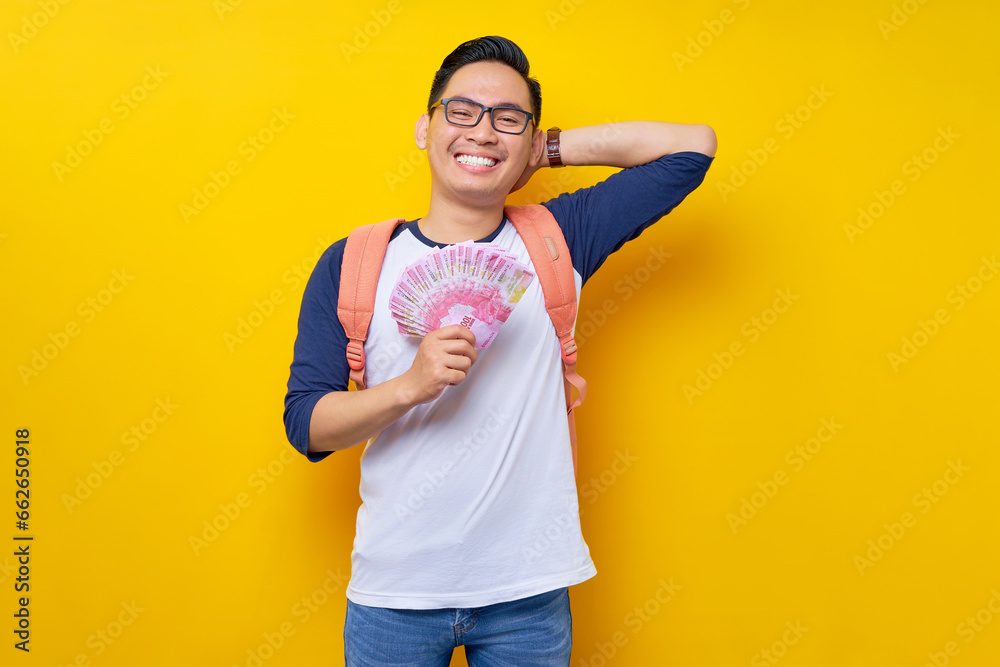 Smiling young Asian student man in casual clothes and glasses backpack holding cash money isolated on yellow background. Education in High School University College concept Smiling young Asian student man in casual clothes and glasses backpack holding cash money isolated on yellow background. Education in High School University College concept