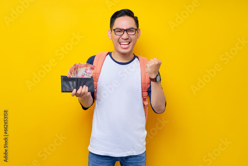 Smiling young Asian student man in casual clothes and glasses backpack holding wallet full of cash money and celebrating winner isolated on yellow background © Bangun Stock Photo