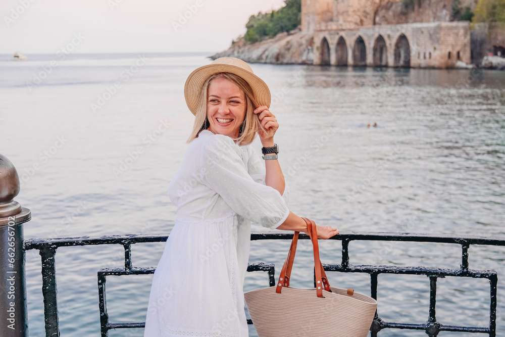 Happy woman in white dress, straw hat and bag laughing. Alanya castle, sea on the background 