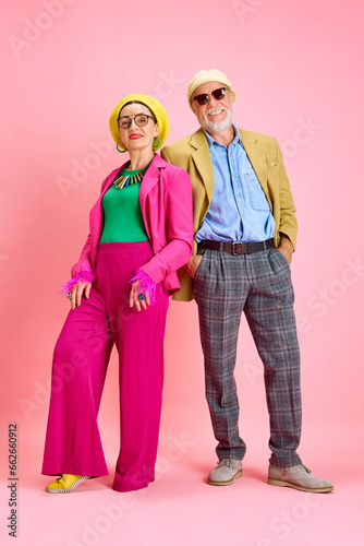 Full-length portrait of beautiful senior couple, elegant man and woman in stylish clothes standing against pink studio background. Concept of beauty and fashion, relationship, modern style, age
