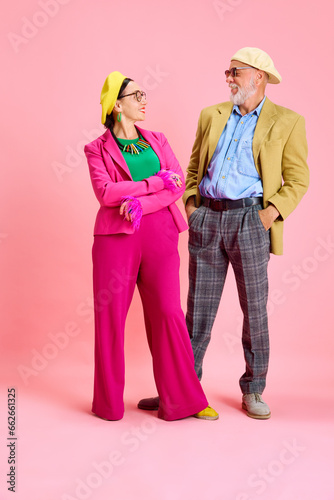 Beautiful elegant senior woman in pink suit and bright accessories looking at happy stylish husband, man against pink studio background. Concept of beauty and fashion, relationship, modern style, age