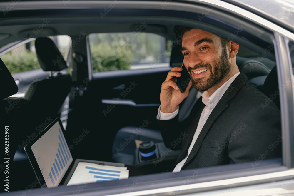 Positive entrepreneur working on laptop and talking on phone while going to airport by car