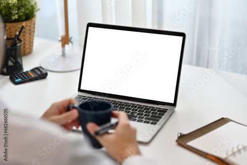 Young female entrepreneur holding coffee cup and using laptop in bright workplace