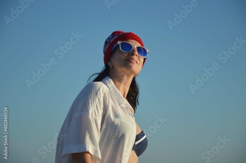 Happy woman wearing a hat with a winter pattern, sunglasses, a white shirt and a swimsuit against the blue sky. Travel and good rest, Christmas holidays, tourism.