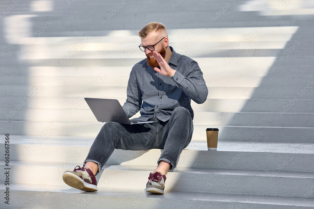 Handsome red-haired freelancer man with a beard sits on the street and works remotely using a laptop