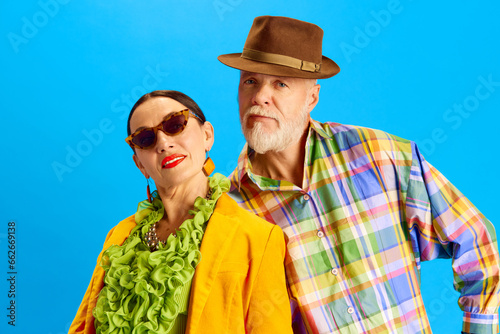 Portrait of beautiful senior couple, attractive woman in sunglasses and bright clothes, handsome man in fedora hat standing against blue background. Concept of fashion, relationship, modern style, age
