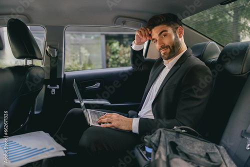 Male sales manager working on laptop sitting on car backseats and looks camera. High quality photo