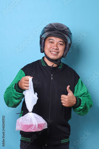 Asian online taxi driver wearing jacket and helmet holding food boxes in plastic bags while showing thumbs up with happy expression