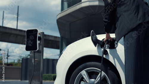 Progressive businesswoman install charger plug from charging station to his electric car before driving around city center. Eco friendly rechargeable car powered by sustainable and clean energy. photo