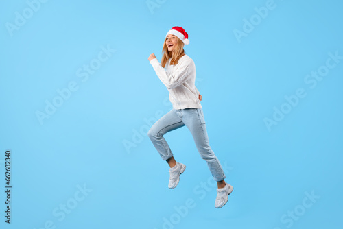 Christmas rush. Happy lady in Santa hat jumping on blue background, excited woman running to buy presents