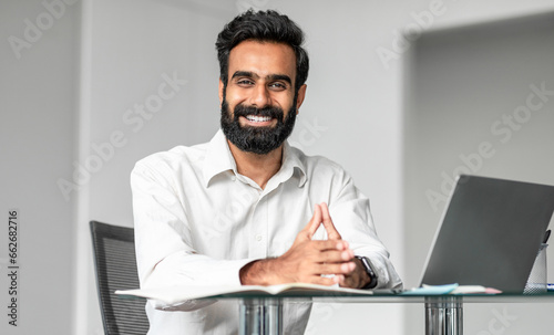 Cheerful confident indian man business consultant working in office, sitting at workdesk in front of laptop and smiling