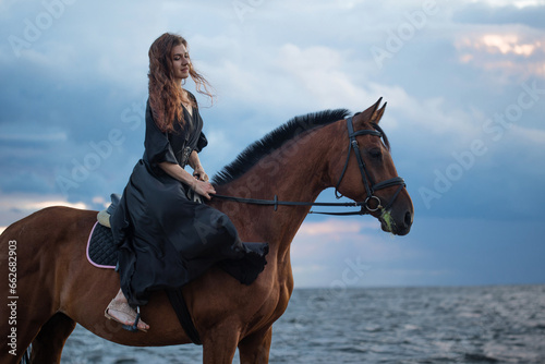 A beautiful young horsewoman in a black dress and with her hair down, riding a horse, portrait against the background of the evening sky, horseback riding in the open air