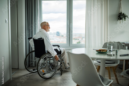 Senior man in a wheelchair spending time alone in apartment. Concept of loneliness and dependence of retired people.