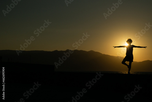 Sagoma femminile di donna riccia, in posizione yoga, con il sole al tramonto alle spalle i cui raggi sfiorano i capelli