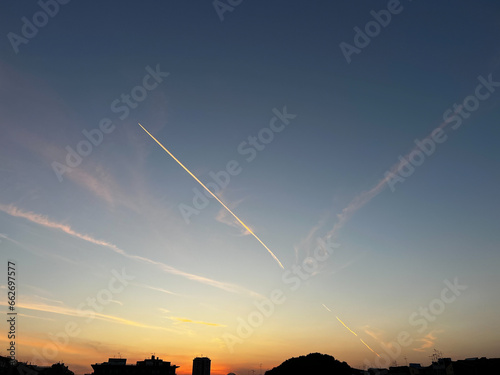 Aereo in volo che lascia una scia di condensazione in un cielo azzurro e arancione al tramonto sui palazzi di Catania photo