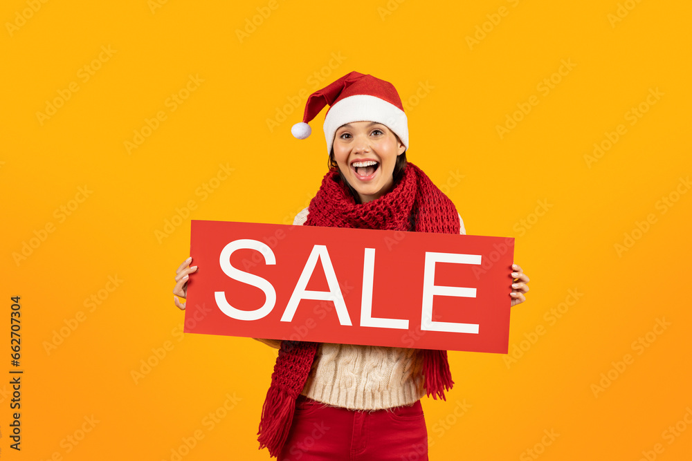 Joyful woman in winter knitwear showing sale poster, yellow backdrop - obrazy, fototapety, plakaty 